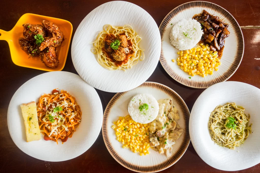 Top view of various plated dishes including chicken wings, spaghetti with meatballs, creamy pasta, garlic bread, grilled pork with rice and corn, and chicken with rice and gravy, served in a casual dining setting.