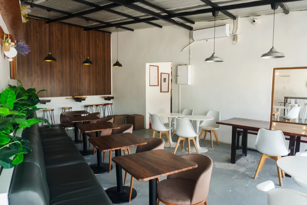 Interior seating area of a coffee shop with a mix of wooden tables, cushioned chairs, and modern white chairs. The space features a wooden accent wall, hanging pendant lights, and potted plants along one side, creating a cozy and minimalist atmosphere